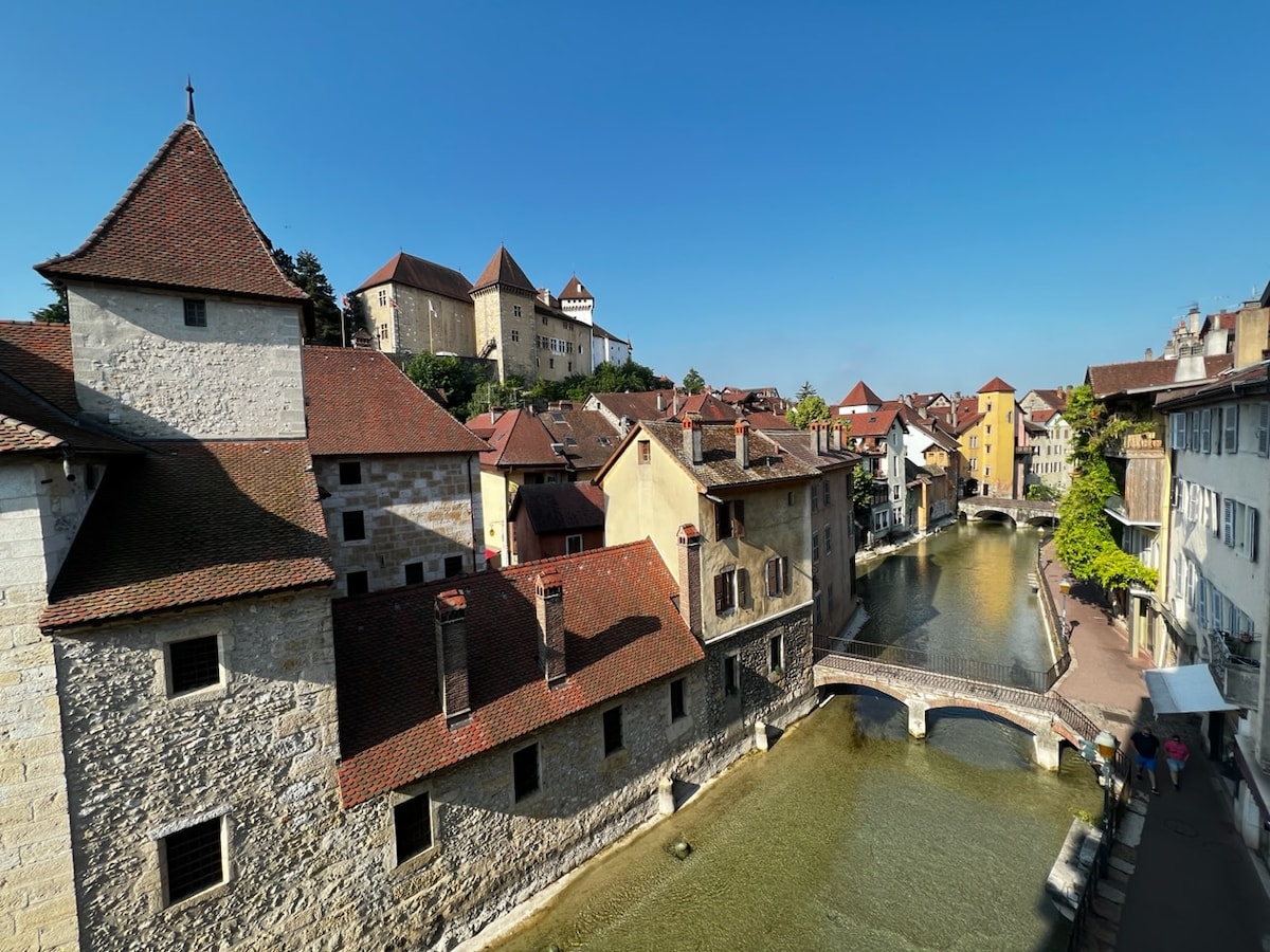 A serene view showcases charming architecture along the canal, with stone and timber houses featuring sloped roofs. The water reflects the vibrant colors of the buildings, while a small stone bridge connects both sides of the canal, enhancing the peaceful atmosphere of the scene.