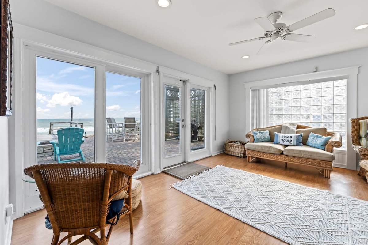 Natural light fills the airy living space, featuring a comfortable seating area with a woven rug underfoot. Large sliding glass doors provide a view of the outdoor patio, which includes seating and a glimpse of the ocean. A fan softly rotates overhead.
