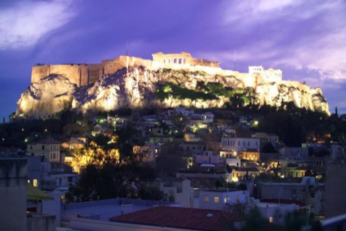 The image captures a panoramic view of the illuminated Acropolis at dusk, with light highlighting its ancient structures. The foreground features a blend of residential buildings, nestled among lush greenery, creating a contrasting backdrop against the historic site.
