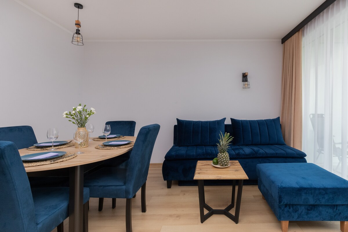 A modern living area is presented with a round dining table set for four, accompanied by elegant blue chairs. A deep blue sofa and pouffe offer comfort, complemented by a wooden coffee table. Natural light filters through sheer curtains, creating an inviting space.