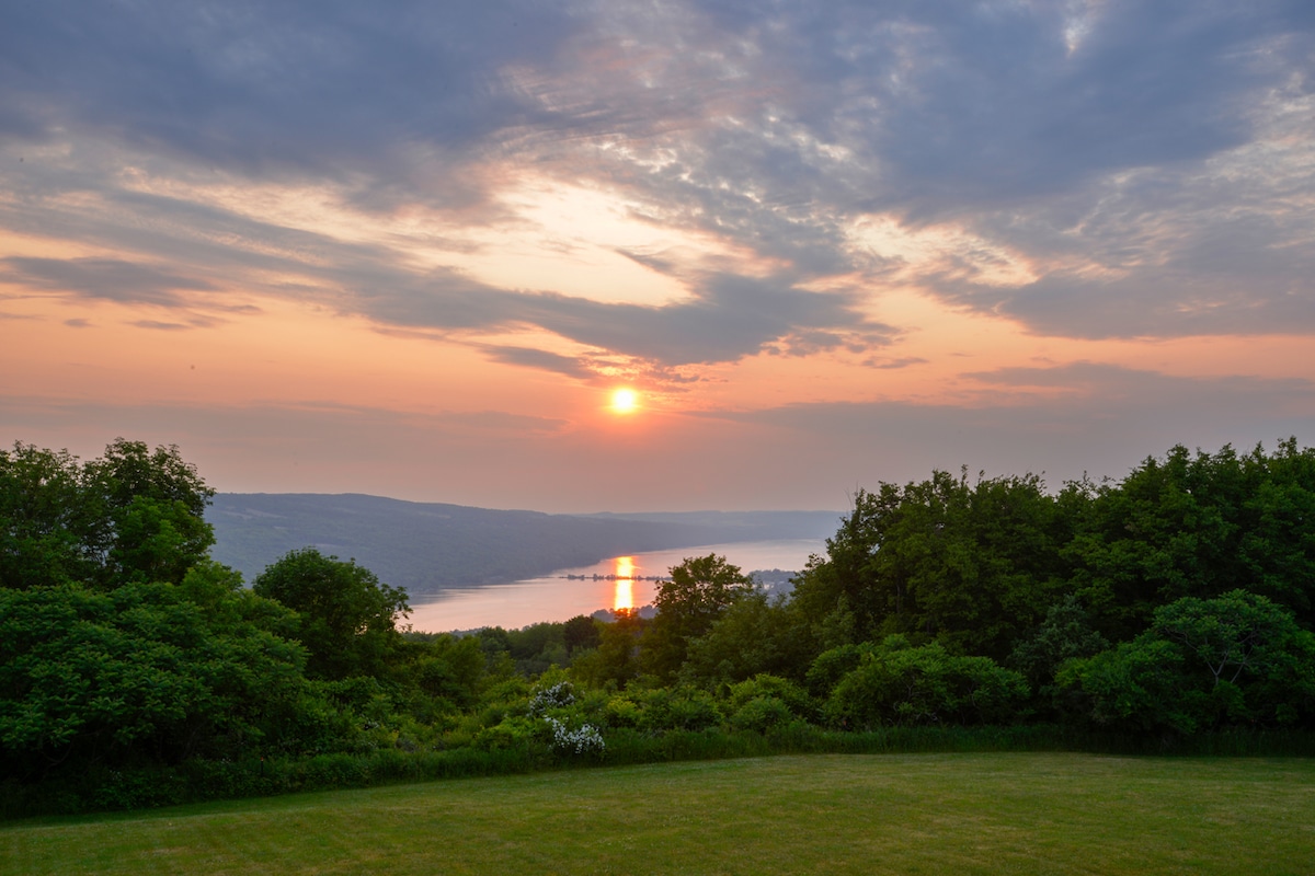 A scenic view of Lake Otisco is captured at sunset, with a blend of orange and purple hues reflected on the water's surface. Lush greenery surrounds the area, creating a serene backdrop against the expansive sky filled with soft clouds.