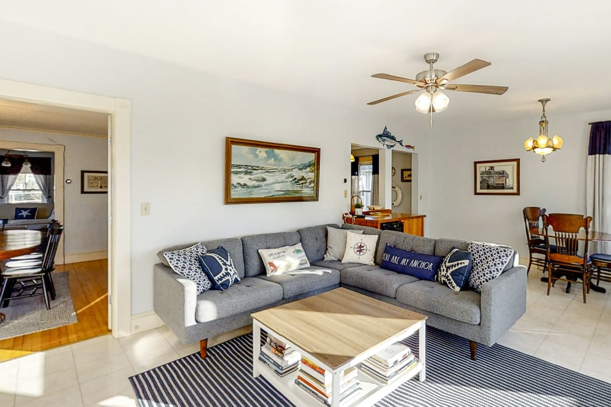 A spacious living area features a modern grey sectional sofa adorned with decorative pillows. A coffee table with an array of books rests on a striped area rug. Wall art, including a landscape painting, adds character, while warm light is provided by a ceiling fan and overhead fixture.