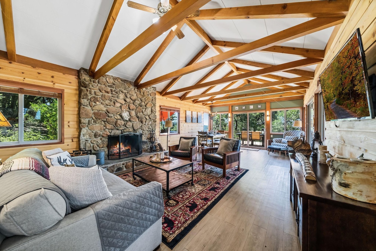 A spacious living area features a large stone wood-burning fireplace, enhancing the warmth of the room. Exposed wooden beams run along the ceiling, and large windows invite natural light. Comfortable seating is arranged around a central coffee table, providing a welcoming space for gatherings.