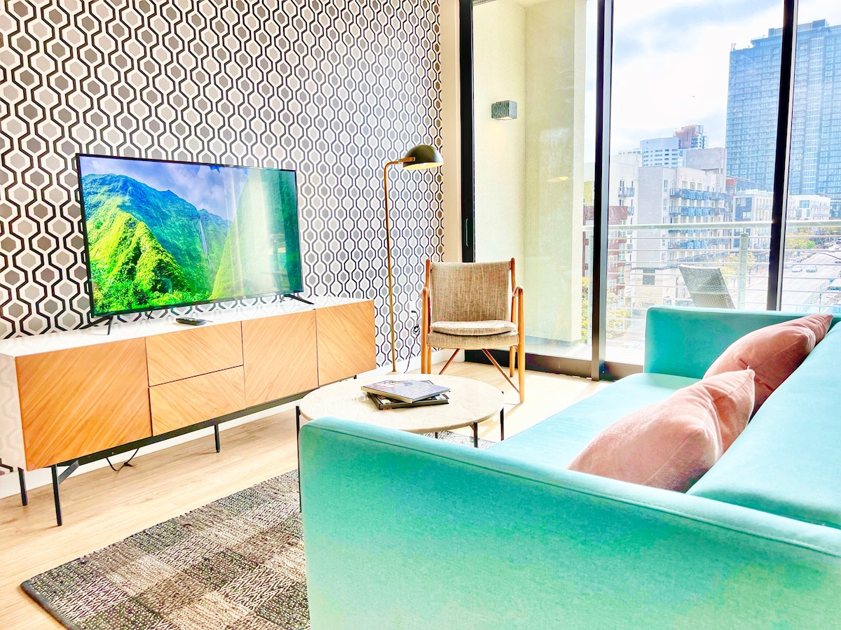 A modern living space features a turquoise sofa complemented by decorative pillows. A coffee table is placed on a textured rug. A wall-mounted television displays a vibrant landscape. Large windows bring in natural light, offering views of the cityscape and creating an open atmosphere.