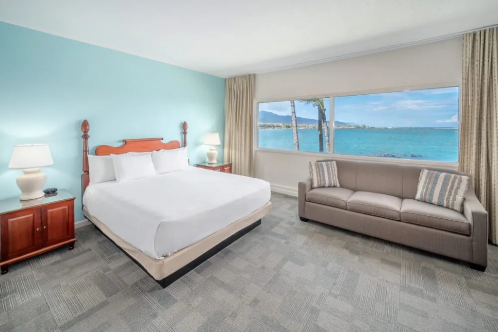 The room features a king bed with crisp white linens and light wood accents, complemented by a modern couch in neutral tones. Large windows provide expansive views of Kahului Bay, allowing natural light to fill the space. A bedside lamp and wooden nightstands complete the inviting setup.