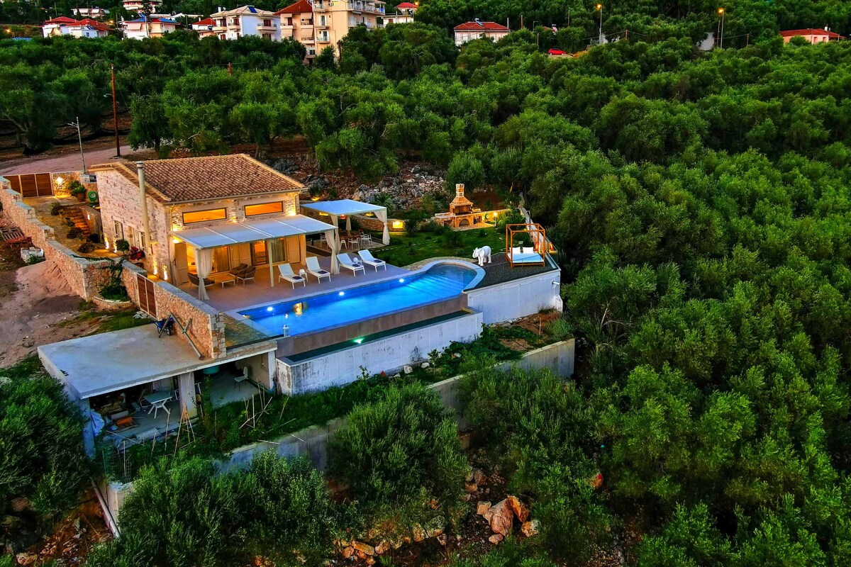An aerial view of the villa showcases a modern design amidst lush greenery. The inviting infinity pool sparkles under ambient lighting, complemented by neatly arranged sun loungers. The tranquil setting is framed by olive trees, with the villa's outdoor terrace extending towards the pool.