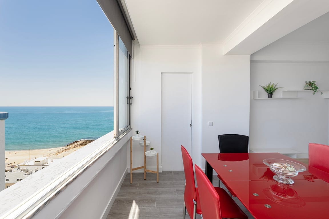 A bright dining area features a red table with four chairs, complemented by a sea view through large windows. A small shelf holds a decorative plant, and a door leads to another room. The beach and ocean are visible in the background.