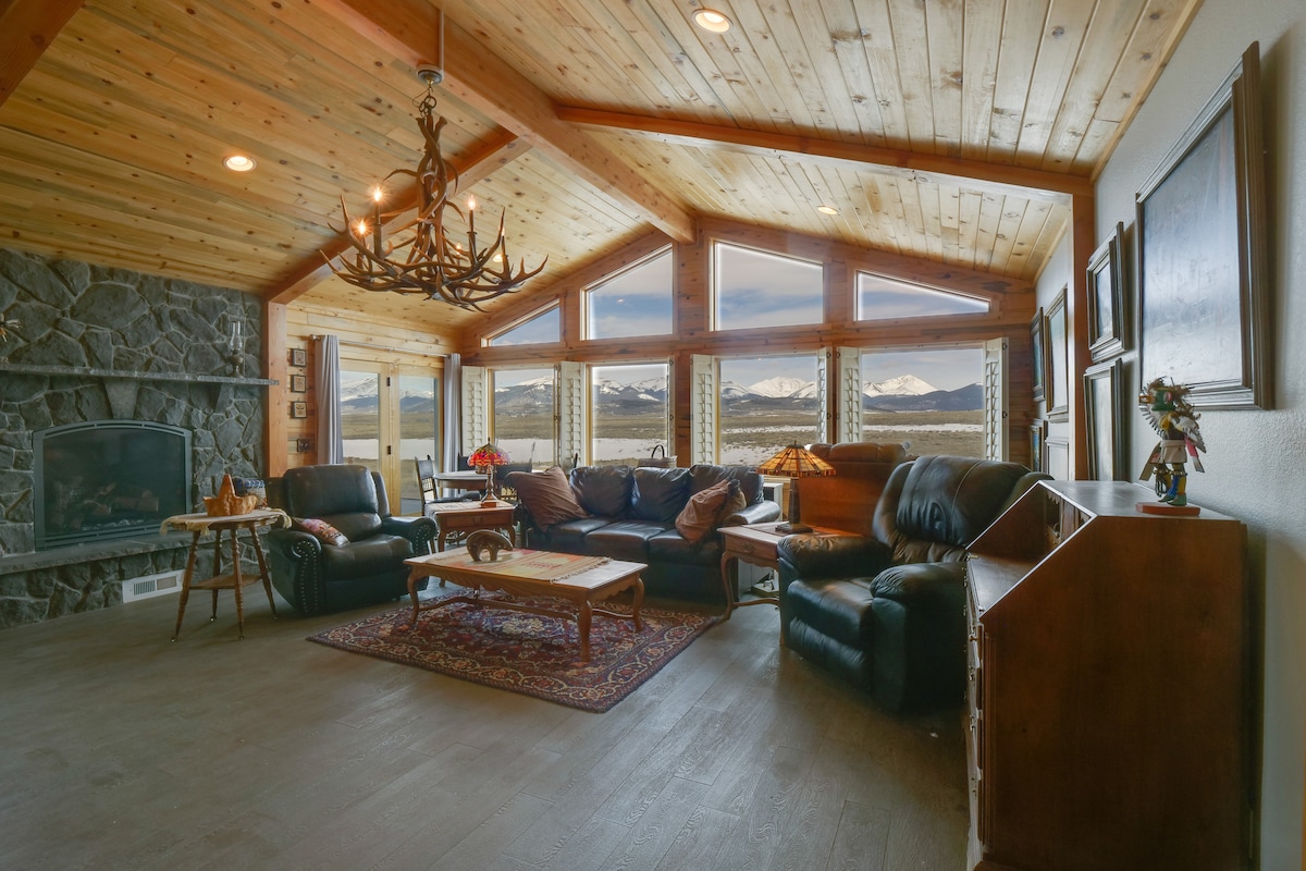 The spacious living area features wooden beams and expansive windows offering stunning mountain views. Comfortable leather sofas are arranged around a central coffee table, complemented by a rustic rug. A stone fireplace adds a cozy touch to the inviting atmosphere.