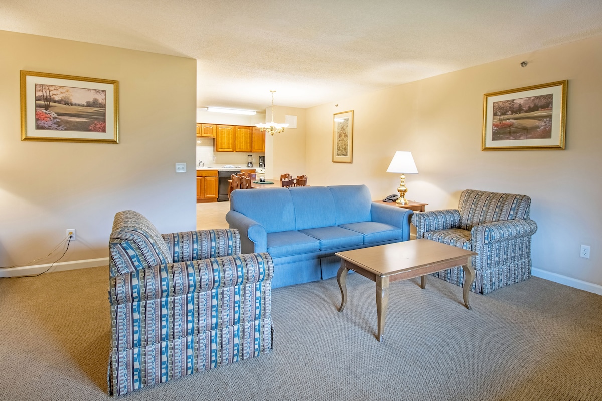 A living area is depicted, featuring a blue sofa and two patterned armchairs arranged around a wooden coffee table. Soft, ambient lighting is provided by a lamp on the table. A doorway leads to the kitchen area, indicating an open floor plan.