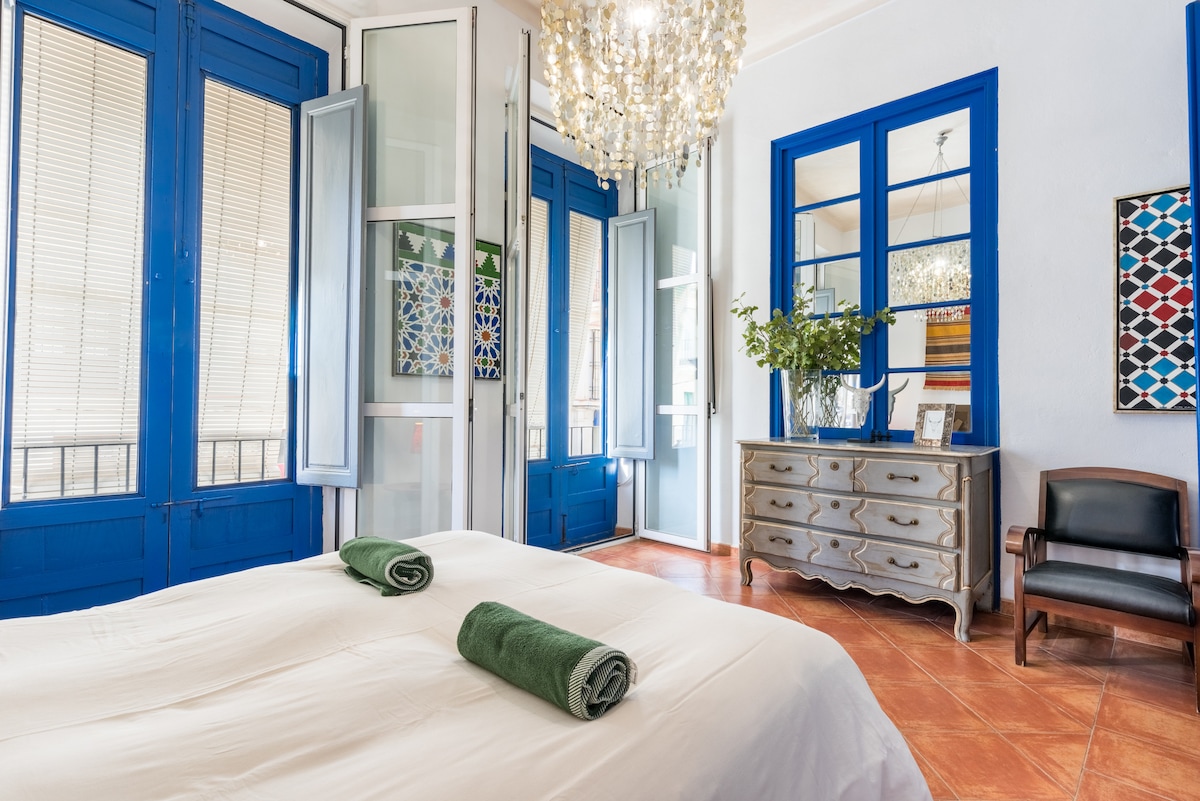 A bright bedroom features a spacious bed adorned with neatly rolled green towels. Large blue-framed windows let in ample natural light and open onto a charming view. A striking chandelier hangs above, while a decorative chest of drawers and a wooden chair add functional elegance to the space.