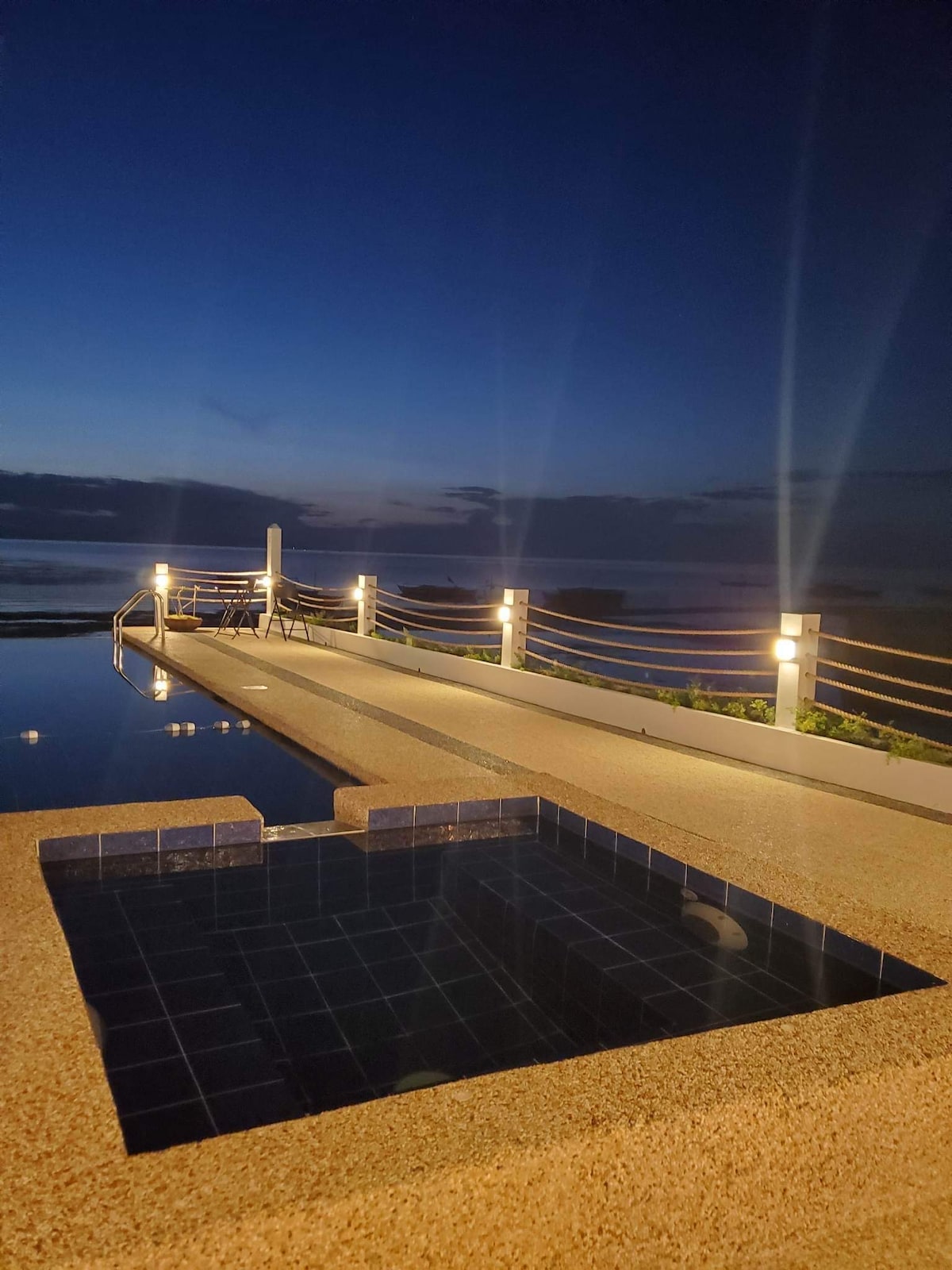 An infinity pool reflects the night sky, illuminated by soft lighting. A small Jacuzzi is positioned at the edge, with wooden railings guiding the path towards the serene sea view. The horizon is visible, blending ocean and sky in twilight.