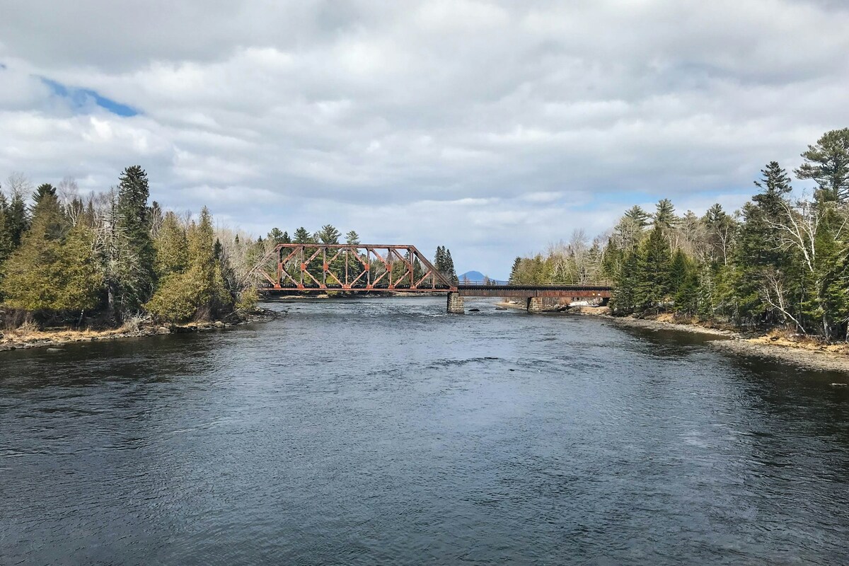 A rustic railway bridge spans the flowing river, framed by lush greenery and trees lining the banks. The calm water reflects the overcast sky, while distant hills create a serene backdrop in the distance, highlighting the natural beauty of the landscape.