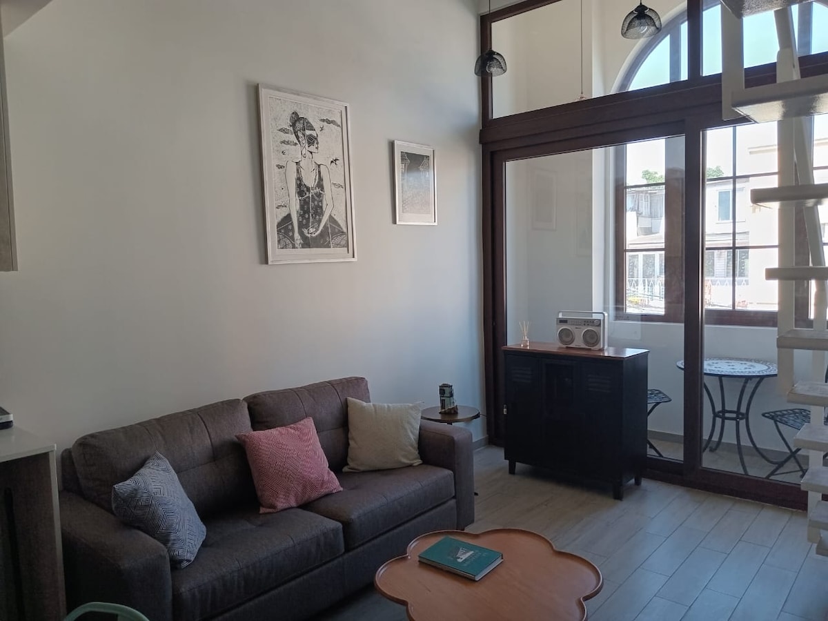 A well-appointed living area features a brown sofa adorned with decorative pillows. A wooden coffee table sits in front, while large windows let in natural light, offering a view of a small balcony with two chairs and a table. Artwork adorns the wall.