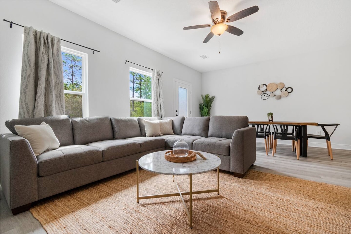 A spacious living area features a plush sectional sofa, complemented by a coffee table atop a natural fiber rug. Large windows invite ample natural light, offering views of the greenery outside. An adjacent dining area includes a stylish table, enhancing the inviting atmosphere.