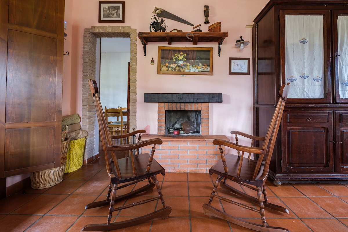 Two wooden rocking chairs are positioned facing a brick fireplace in a warmly colored living space. Shelves adorned with rustic decor and a framed painting are mounted on the walls. Traditional cabinets and a doorway leading to another room are visible in the background.