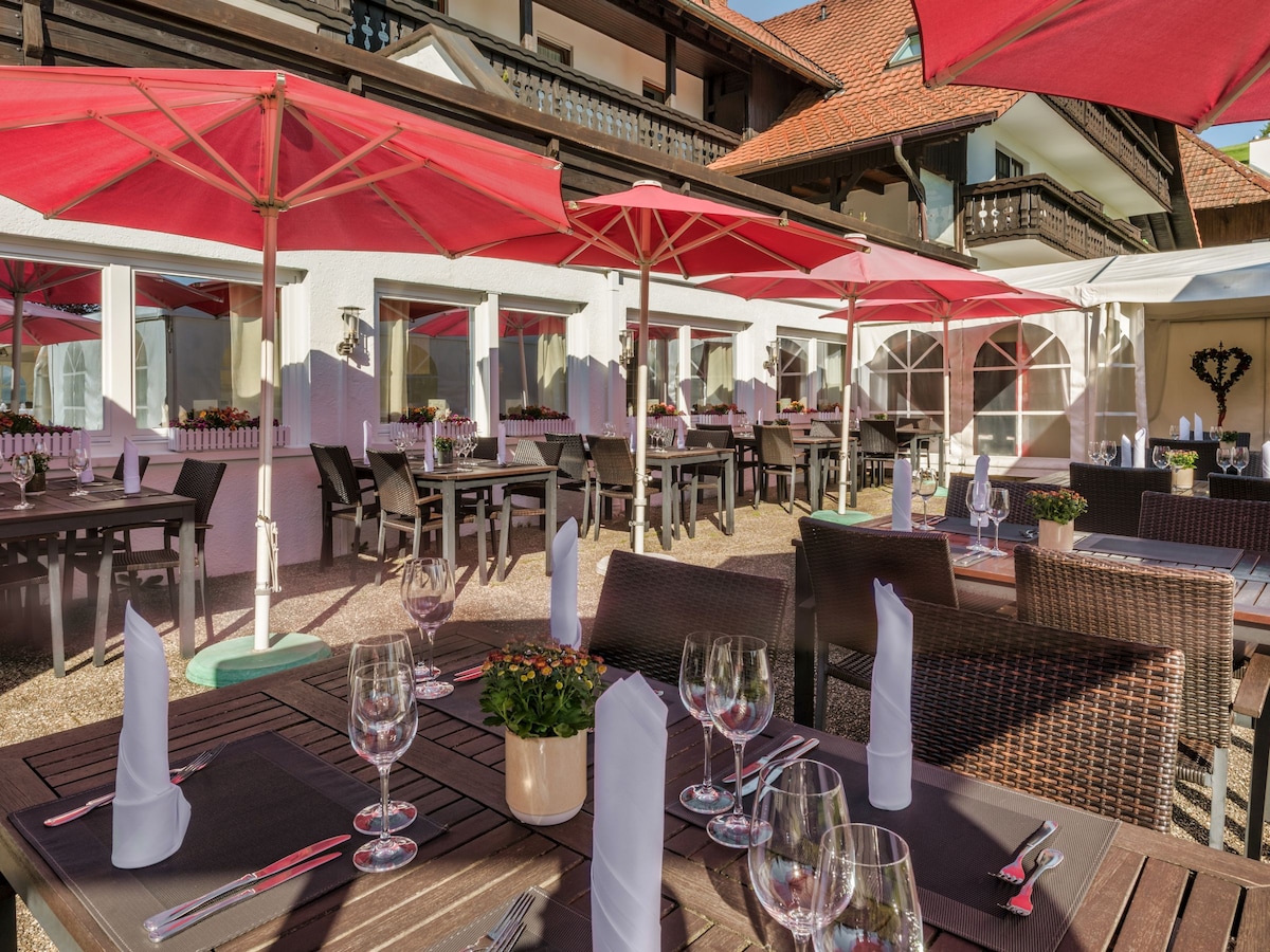 An outdoor dining area features several tables set with glassware and neatly arranged cutlery. Large red umbrellas provide shade for the guests, with floral centerpieces enhancing the setting. Surrounding architecture showcases traditional wooden elements and open balconies, suggesting a welcoming atmosphere.