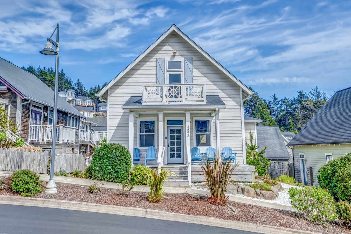 A charming coastal home is presented with a light-colored exterior, featuring a welcoming porch adorned with blue Adirondack chairs. Lush greenery and a landscaped yard frame the entrance, while the upper balcony offers additional outdoor space under a clear blue sky.