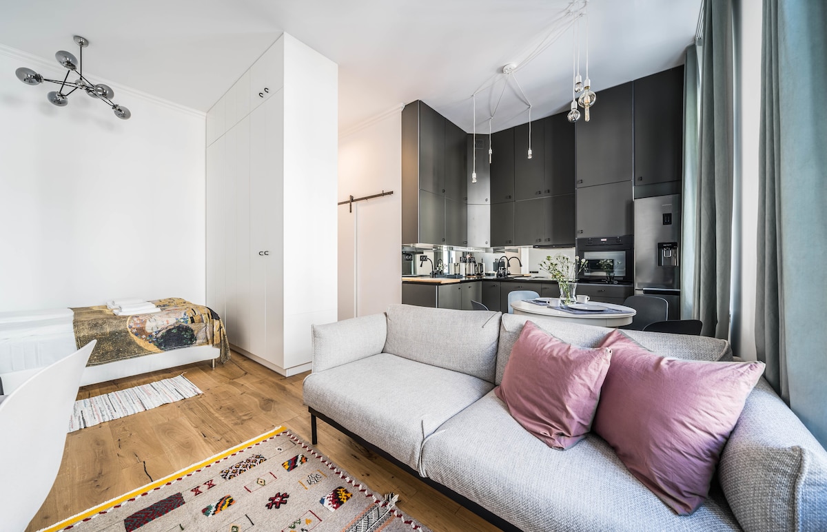 A modern studio living space is shown, featuring a comfortable sofa with pink cushions and a dining area. A fully equipped kitchenette is visible in the background, while light streams through large windows dressed in soft curtains. A cozy sleeping area is indicated by a partially visible bed.