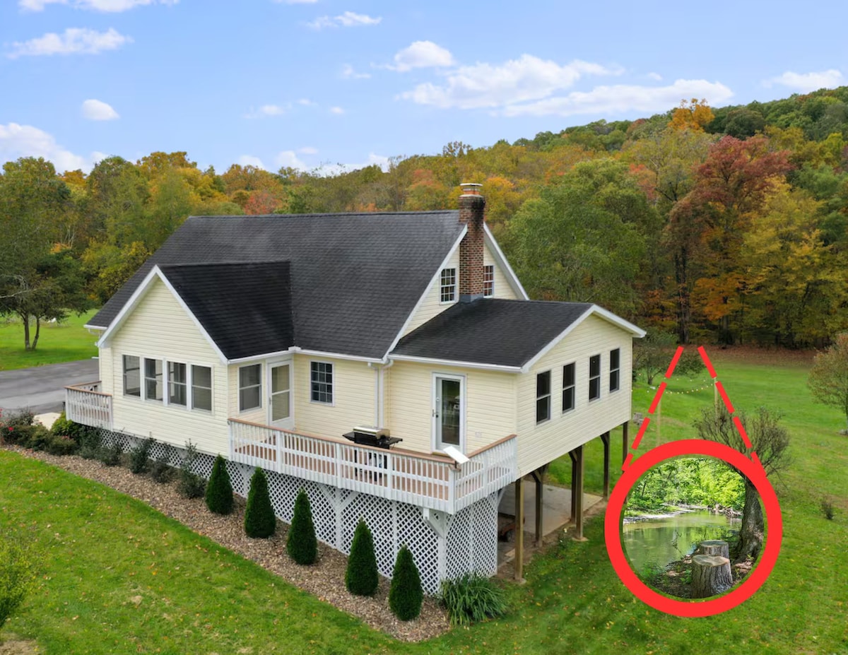 The exterior of the house is shown, featuring a charming light-colored facade and a wraparound deck. The surrounding landscape includes lush greenery and colorful fall foliage. In the foreground, a serene creek can be seen, enhancing the natural beauty of the location.