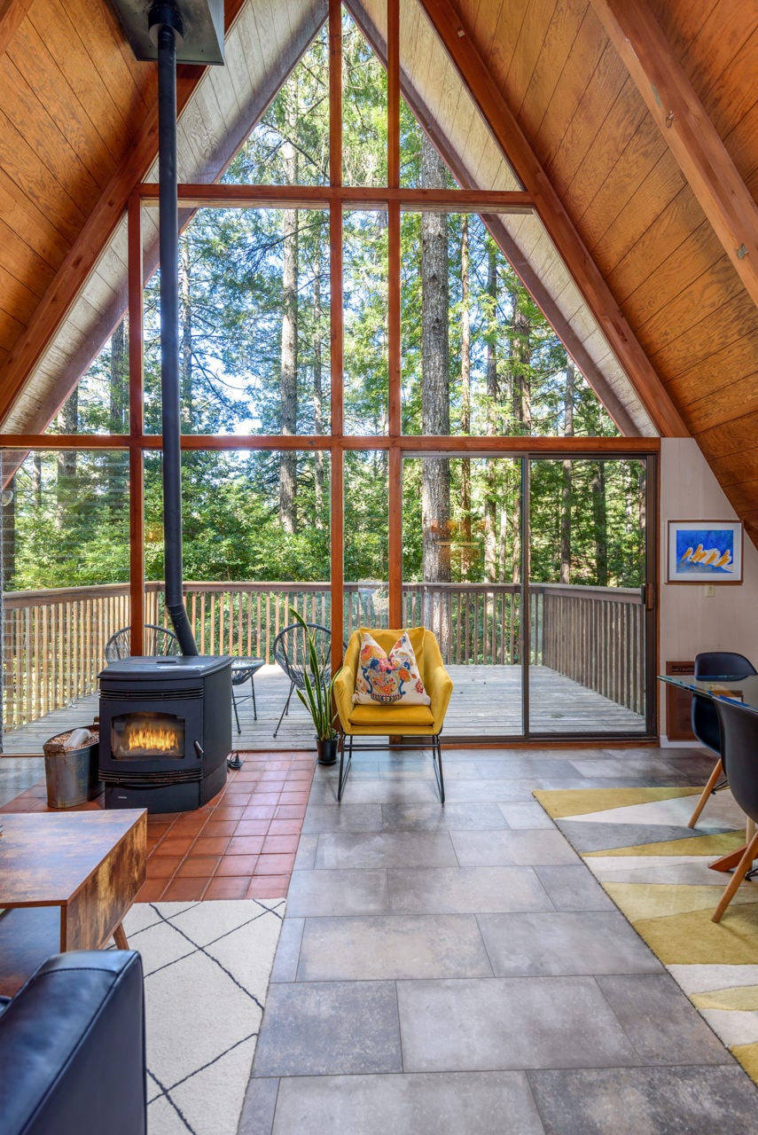 A cozy living area features an A-frame structure with floor-to-ceiling windows that frame vibrant trees outside. A wood-pellet burning stove adds warmth, while a yellow chair and patterned rug provide accents. Sliding doors lead to a spacious deck surrounded by nature.