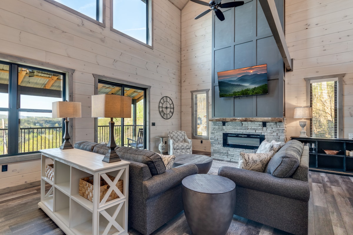 A spacious living area features large windows that provide views of the surrounding landscape. Comfortable seating options are arranged around a central table, with a large television mounted above an elegant stone fireplace. Natural light fills the room, creating a bright and inviting space.