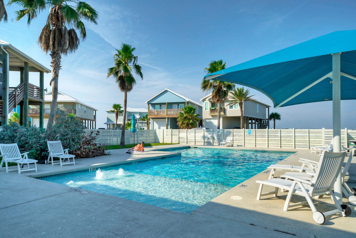 A community pool is surrounded by lounge chairs and palm trees. Sunlight reflects off the water, and a shaded area offers respite from the sun. Nearby, well-kept homes provide a coastal backdrop, enhancing the relaxed atmosphere of the outdoor space.