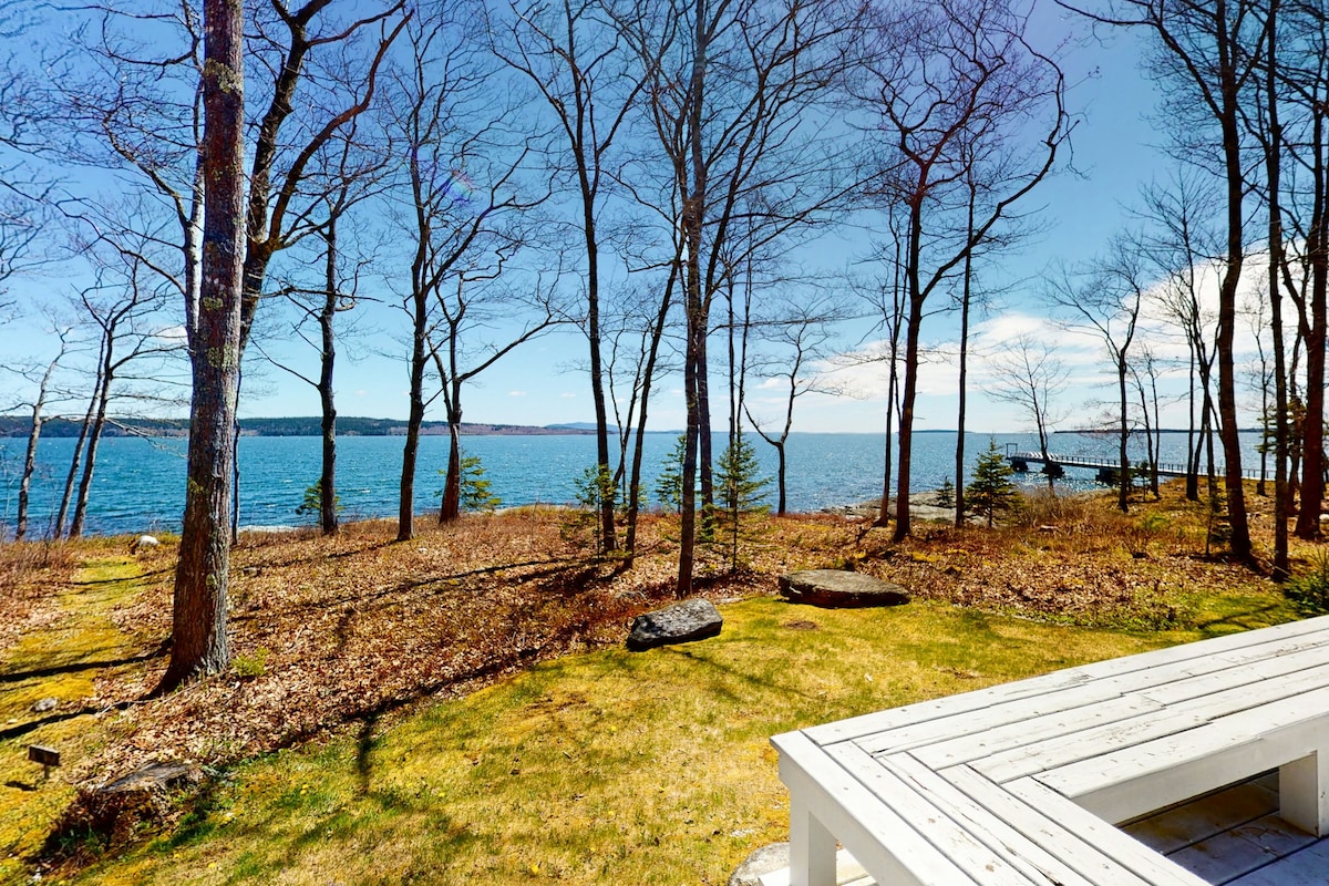 A scenic view of the ocean can be seen through the trees, with vibrant blue water meeting the sky. A wooden deck is positioned in the foreground, providing a space for outdoor seating. The natural landscape includes patches of grass and rocky elements.