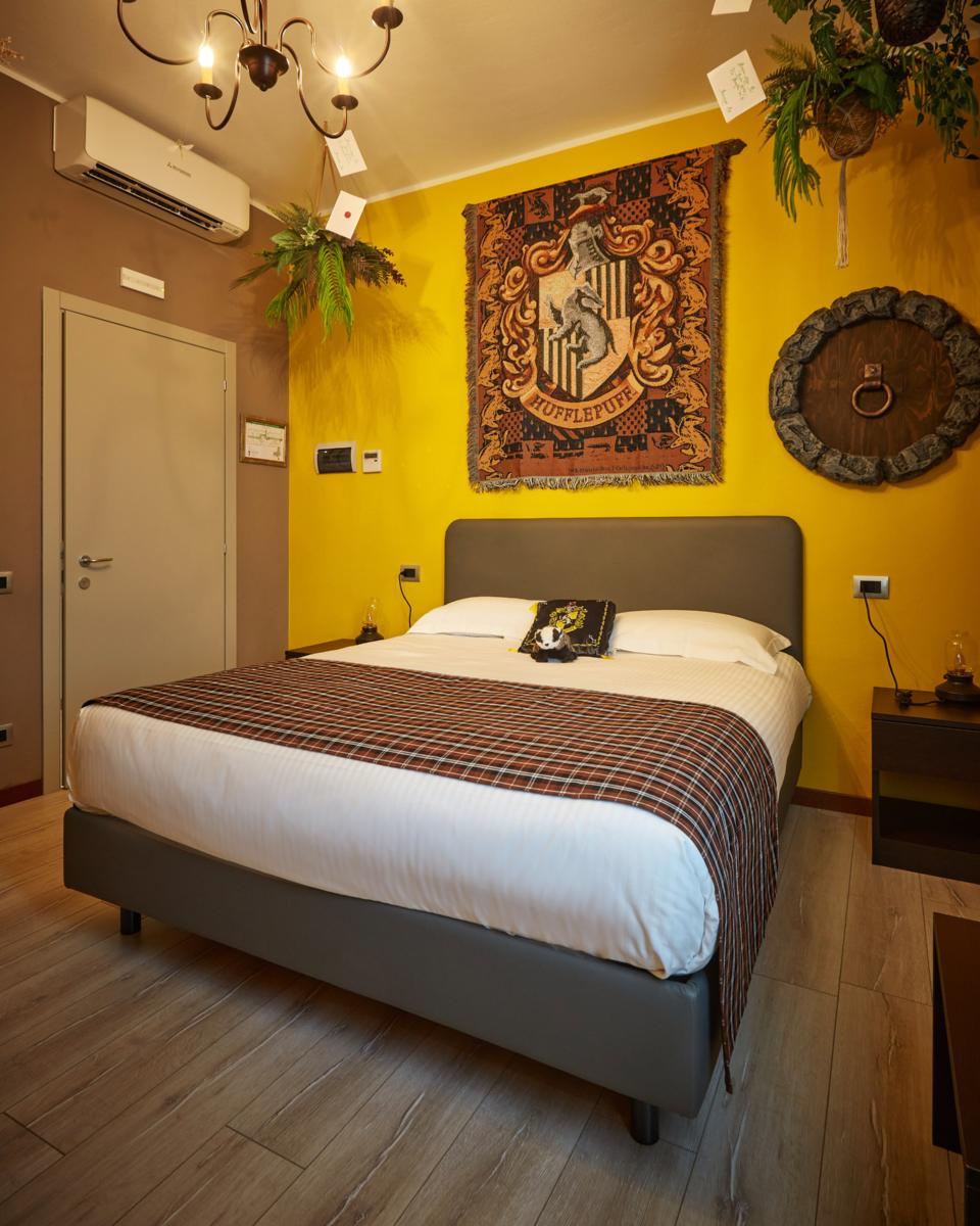 A cozy queen-size bed is positioned against a vibrant yellow wall, accompanied by a striking tapestry featuring the Hufflepuff emblem. Decorative plants are suspended from the ceiling, and a nightstand with a small decorative item is placed beside the bed. The room is lit by a stylish chandelier.