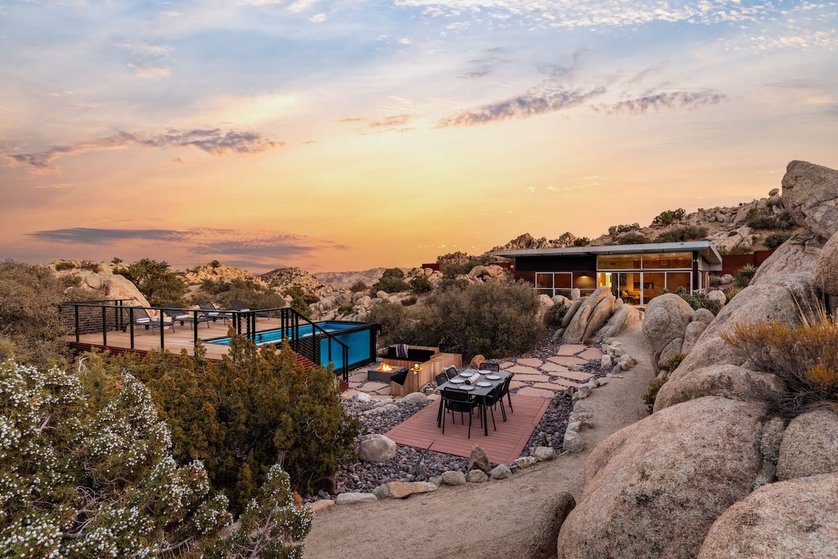 The exterior view features a modern retreat set against a desert sunset. The house blends with the landscape, surrounded by boulders and desert vegetation. An inviting pool area is visible, complemented by outdoor seating arrangements, showcasing the seamless integration of architecture and nature.