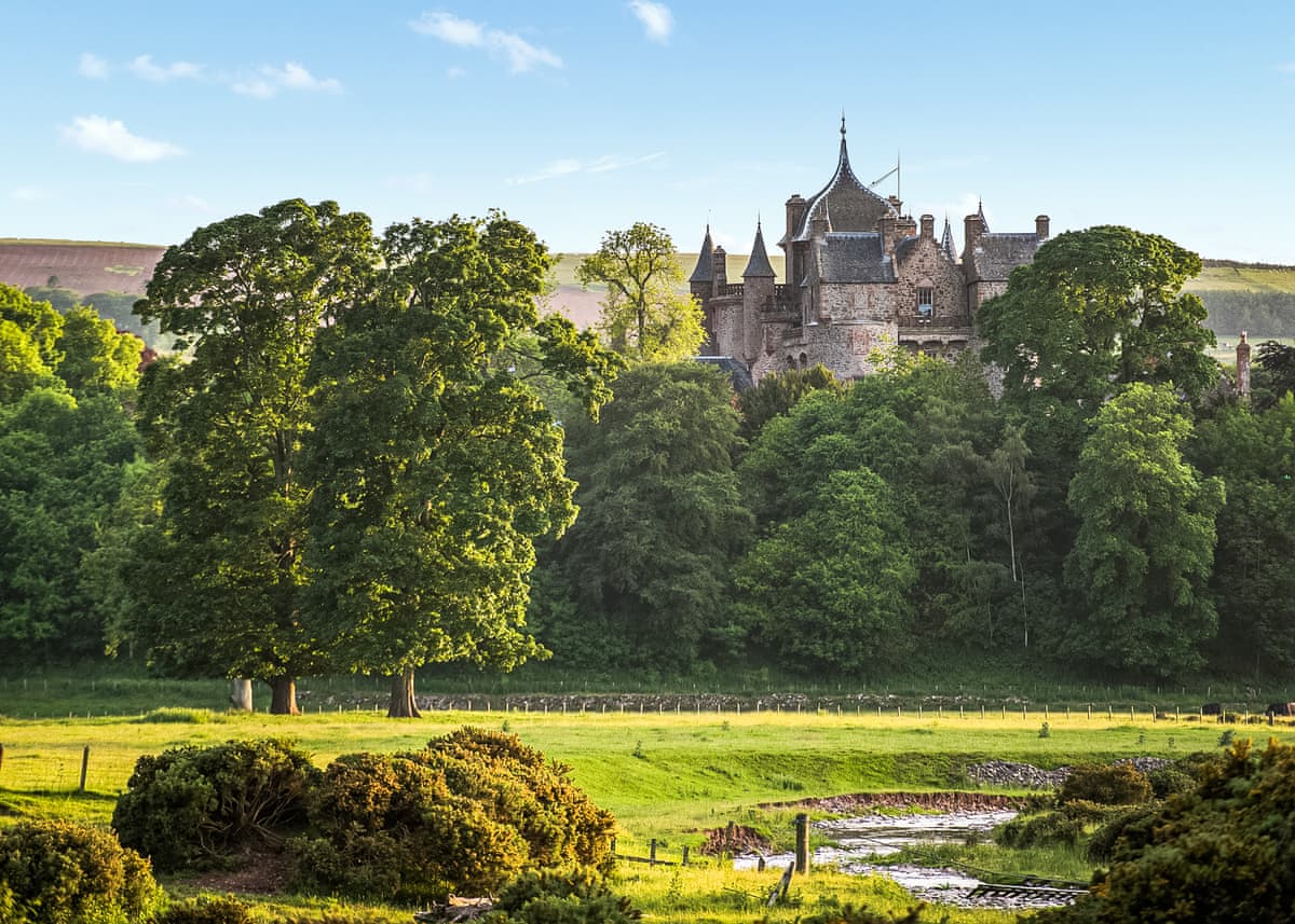 A historic castle is nestled among lush greenery, partially obscured by trees. The structure features turrets and intricate architectural details, with a serene landscape surrounding it. A gentle river flows in the foreground, and rolling hills rise in the background under a clear blue sky.