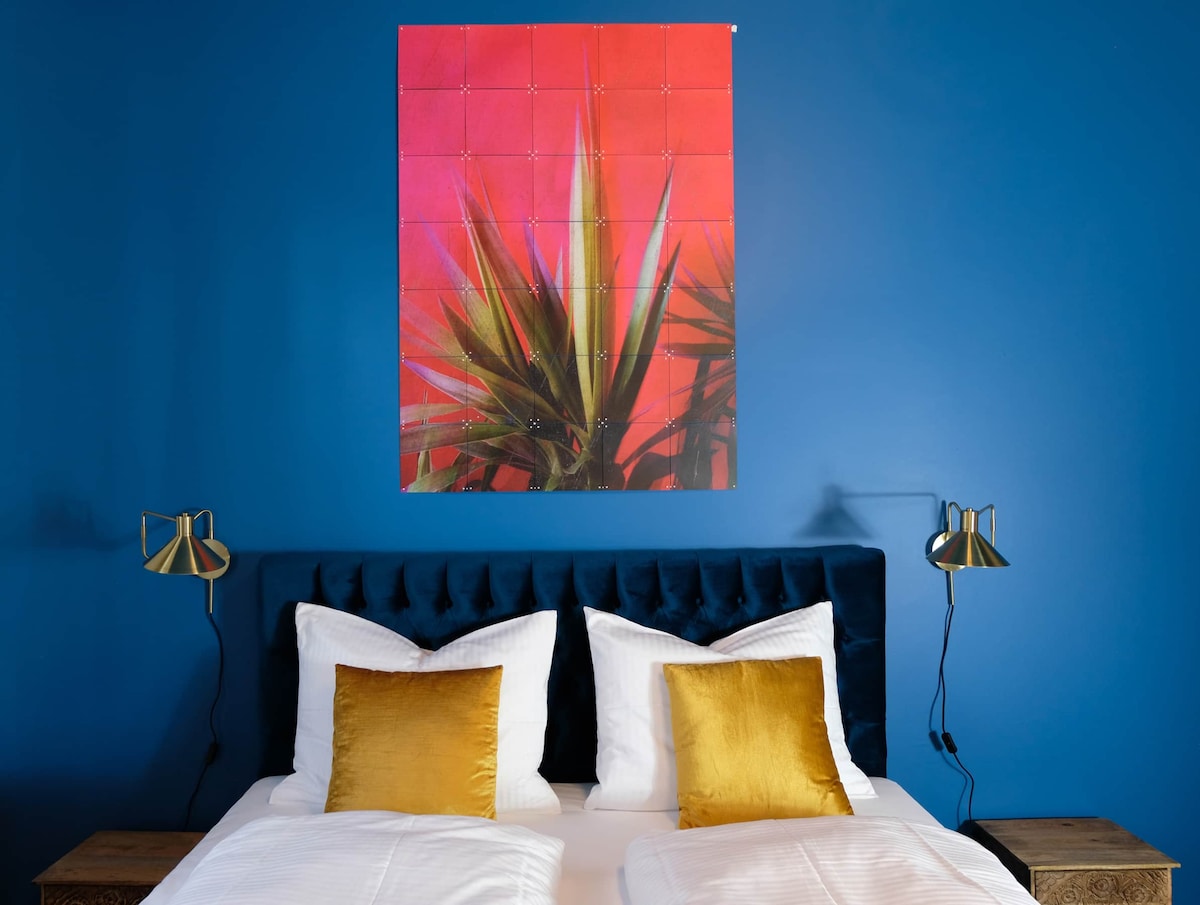 A spacious bed is dressed in white linens, accented by two golden cushions. A vibrant red and green wall art piece depicting plant life hangs above the bed. Stylish bedside lamps with unique designs are positioned on either side.