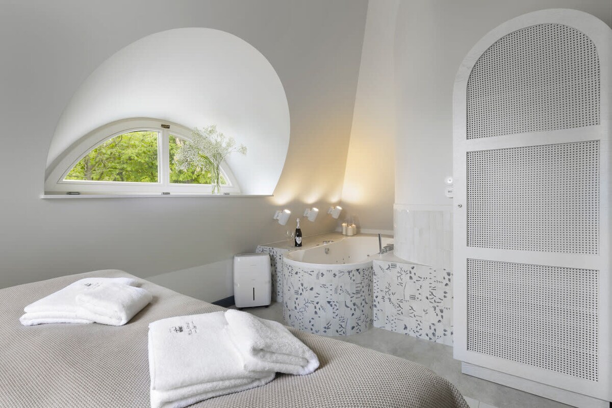 A bright and serene bedroom features an arched window allowing natural light to enhance the space. A spacious jacuzzi is set against a decorative wall, complemented by soft towels positioned nearby. The elegant décor is characterized by muted tones and modern design.