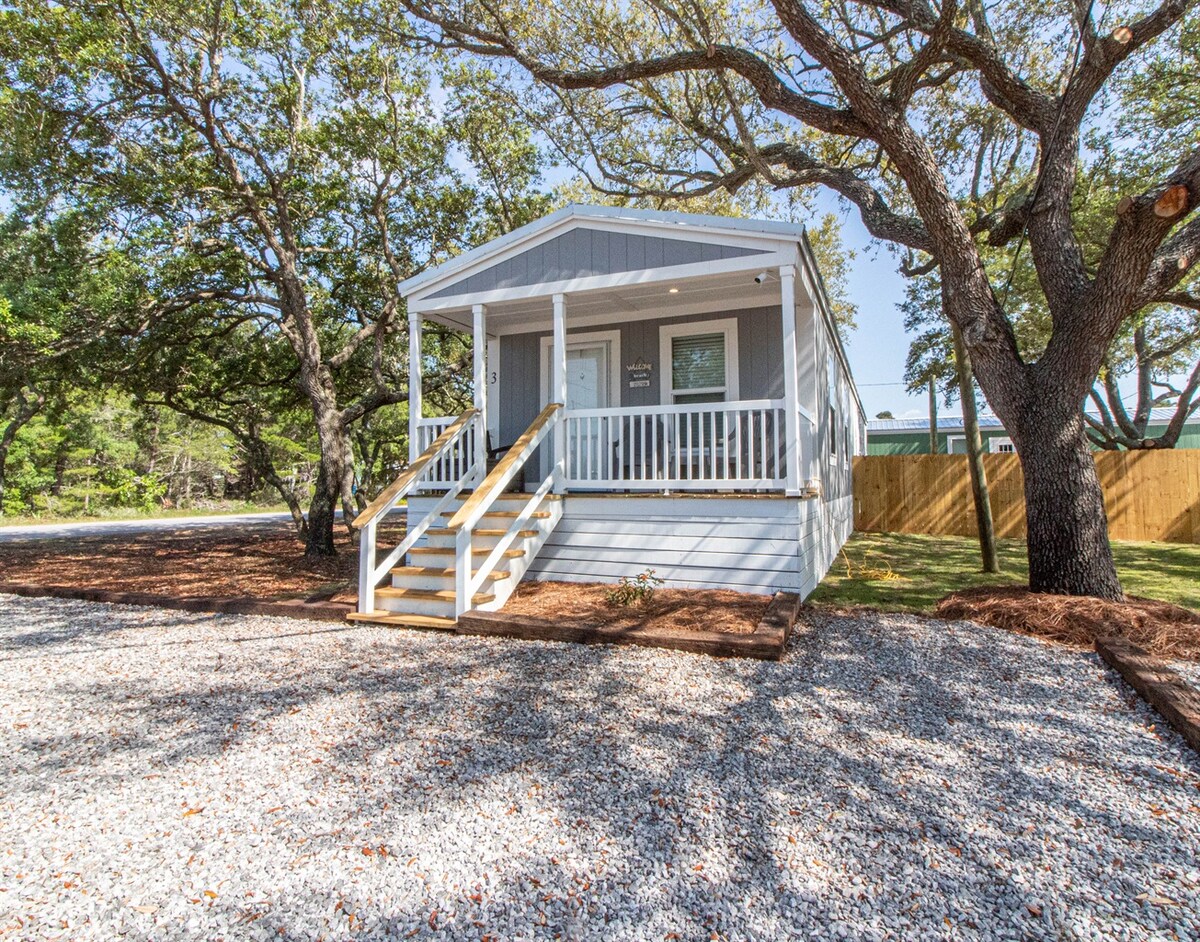 A charming cottage is shown with a covered porch and white railings. A gravel pathway leads up to the entrance, surrounded by landscaped areas with trees and shrubs. Ample outdoor space is available on the property, adding to the inviting exterior.