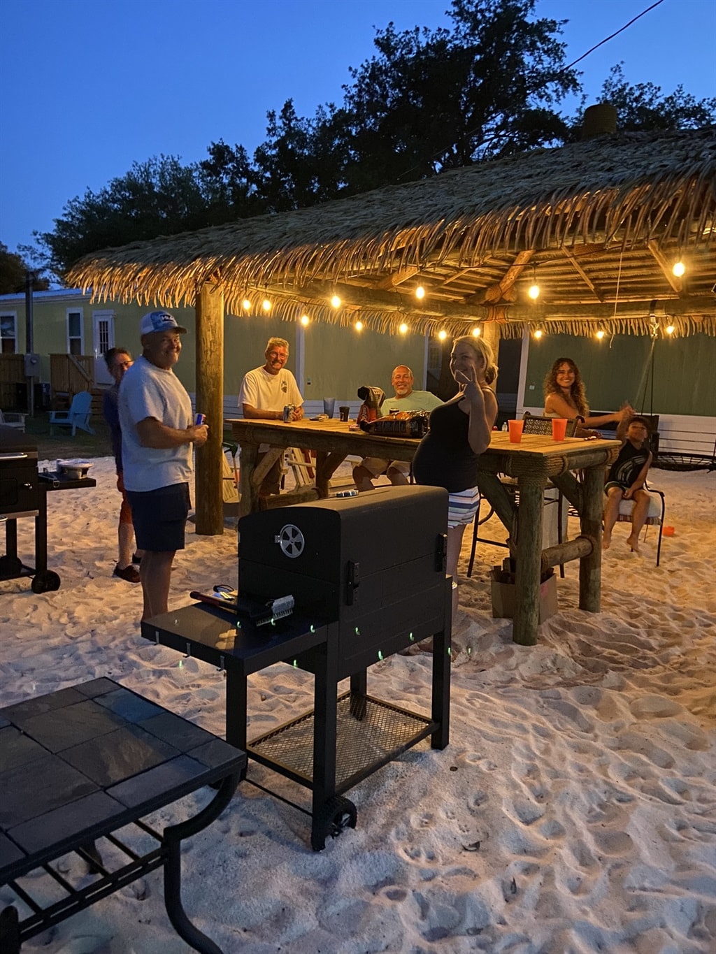 An outdoor communal area features a large wooden table with string lights overhead, creating a relaxed setting under the evening sky. Grill stations are placed nearby on a sandy surface, surrounded by several seated individuals enjoying their time together.