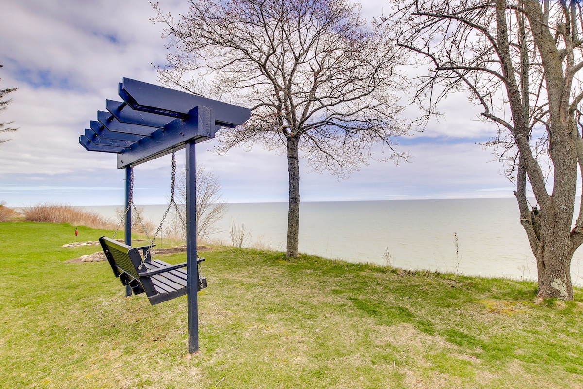 A bench swing is positioned under a wooden pergola, overlooking a calm lake. Lush green grass surrounds the area, while bare trees provide a natural backdrop. Soft clouds linger in the sky, reflecting the tranquil mood of the lakeside setting.