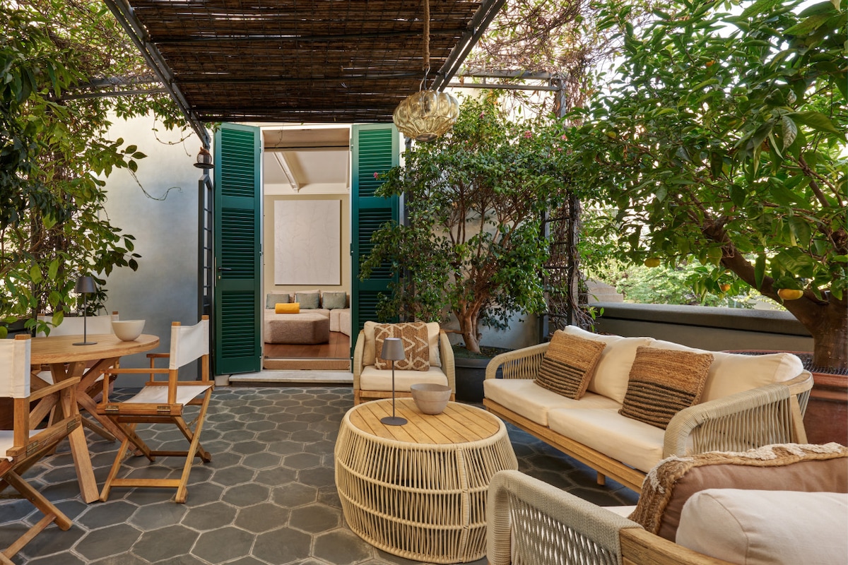A private terrace features a light wooden dining table and comfortable seating areas surrounded by lush greenery. The space is enhanced by the presence of decorative elements, such as a woven pendant light, creating a tranquil outdoor setting accessible from the living area.
