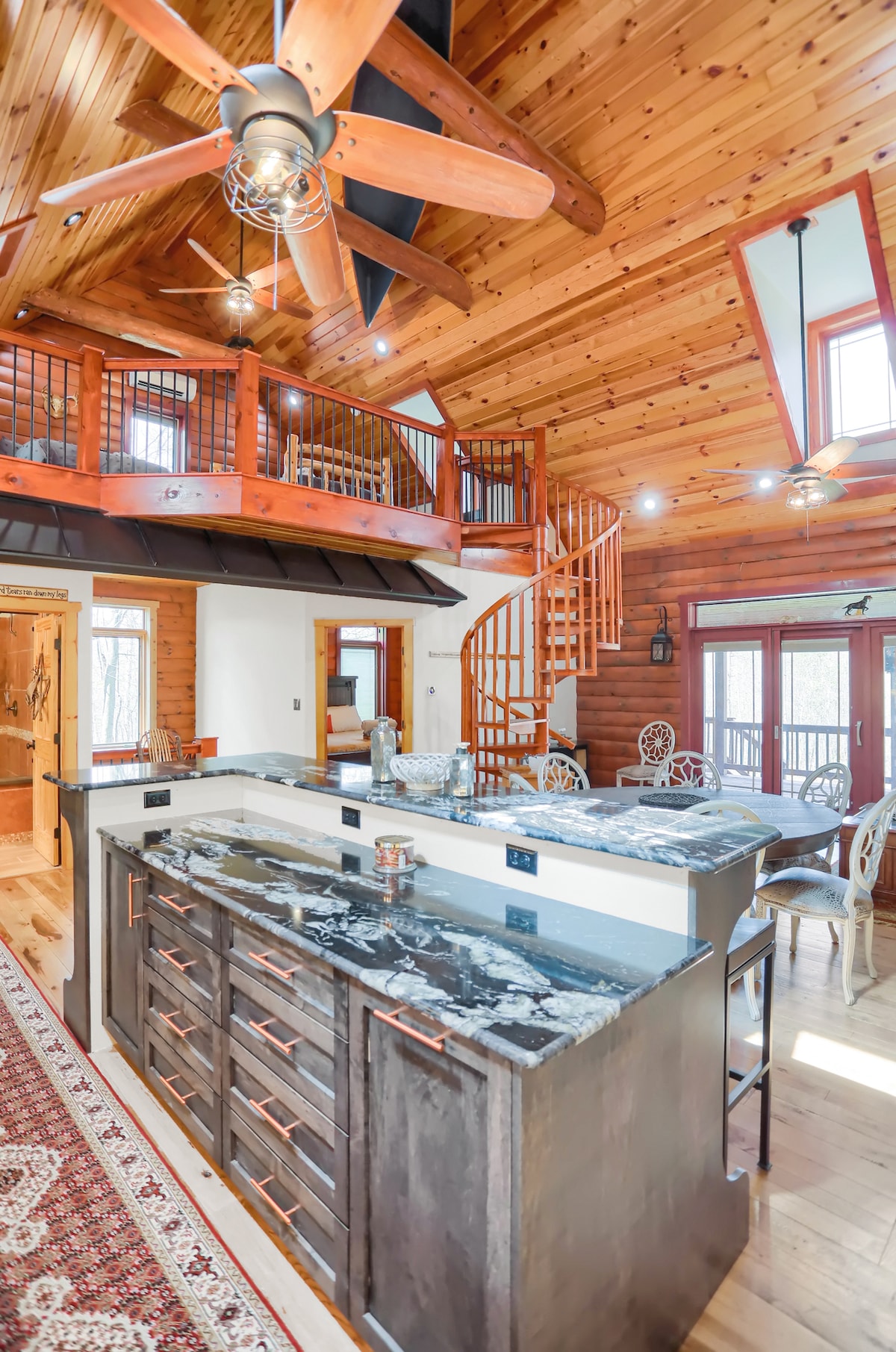 An open kitchen features dark wooden cabinetry and black granite countertops, providing a modern touch. A spiral staircase leads to a lofted space, while wooden beams and large windows allow for ample natural light. A dining area with intricate chairs is visible, enhancing the inviting layout.