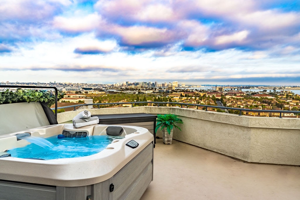 A luxurious jacuzzi is situated on a private deck, surrounded by a stunning panoramic view of the bay and city skyline. Soft clouds float above, enhancing the serene atmosphere. A potted plant provides a touch of greenery, complementing the inviting setting.