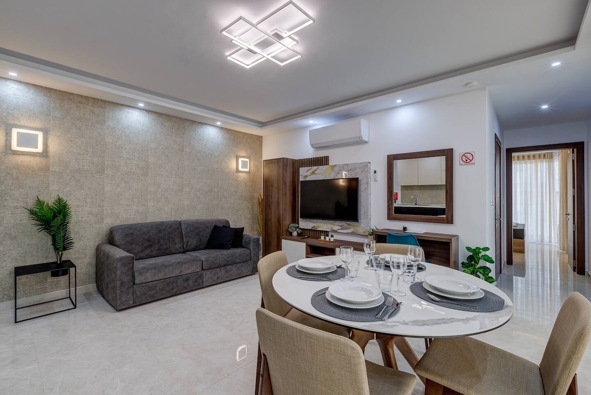 The living area features a modern, grey sofa set against a textured wall, complemented by a stylish dining table with place settings. A flat-screen TV is mounted on the wall, and natural light filters in through a nearby window, enhancing the inviting atmosphere.