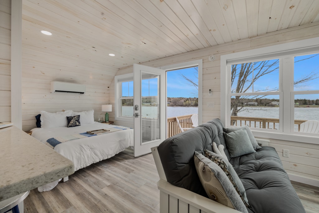 A bright and airy interior features a cozy sleeping area with a spacious bed, while a comfortable sofa sits nearby. Large windows provide views of the surrounding landscape, and double doors open to an outdoor space, inviting natural light into the room.