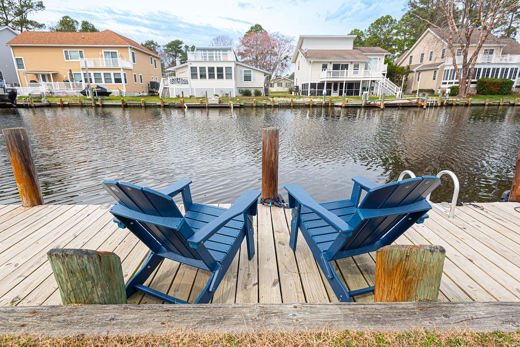 Two blue Adirondack chairs are positioned on a wooden dock, facing the calm canal waters. The dock is framed by wooden posts, with residential homes visible across the waterway, creating a serene waterfront setting.