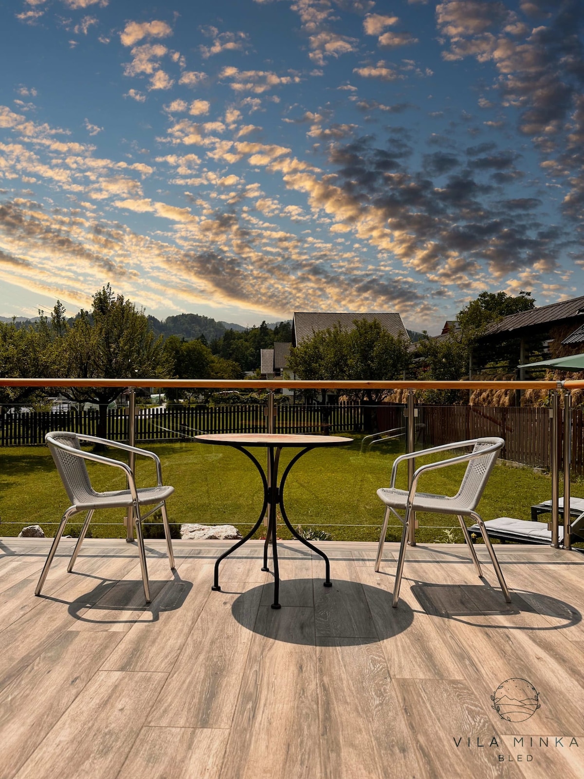 A terrace features a small round table and two modern metal chairs, positioned on a wooden deck. Lush green grass extends beyond the terrace, framed by trees and a wooden fence against a backdrop of a colorful sky with scattered clouds.