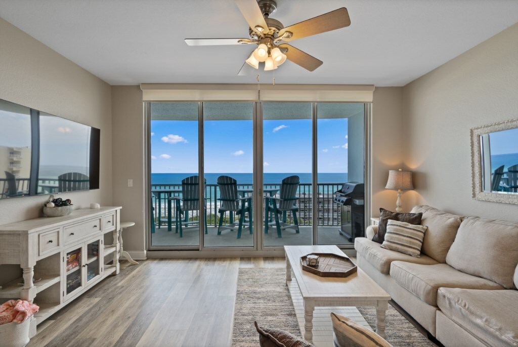 A bright living area features large glass doors that open to a Gulf-view balcony. A comfortable sofa and a neutral-toned coffee table are positioned on a light wooden floor, while a ceiling fan adds to the airy ambiance. A mirrored wall reflects the natural light.