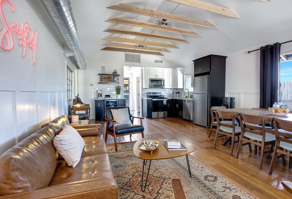 A spacious living area features wooden beams on the ceiling, complemented by a cozy leather sofa and a decorative rug. The open kitchen is equipped with modern appliances, including a refrigerator and oven, while a dining table with chairs is positioned nearby for shared meals.