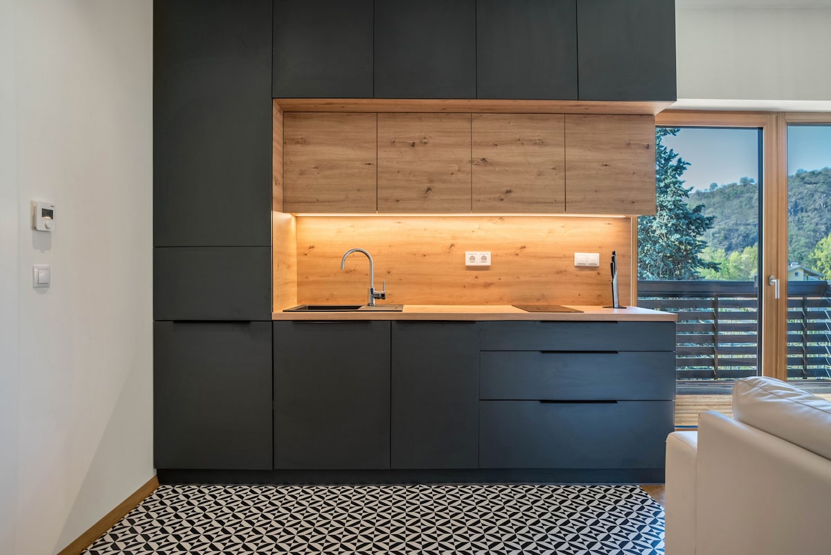 A modern kitchen area is featured, with sleek black cabinetry and a wooden countertop. Under-cabinet lighting highlights the workspace, while a sink and faucet are positioned against the wall. A patterned tile floor adds visual interest, complementing the overall design.