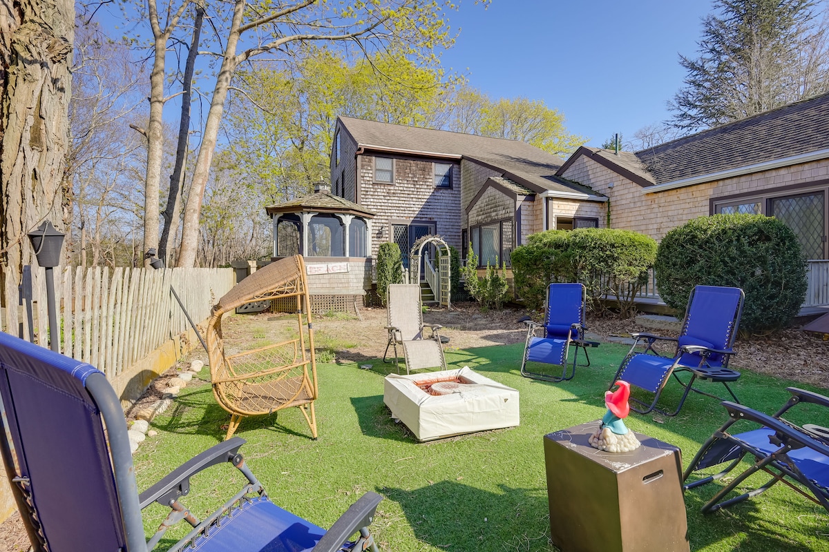 A spacious outdoor area features landscaped grass, surrounded by trees and a wooden fence. Comfortable lounge chairs are arranged around a fire pit, and a screened gazebo is visible in the background, providing a shaded spot for relaxation.
