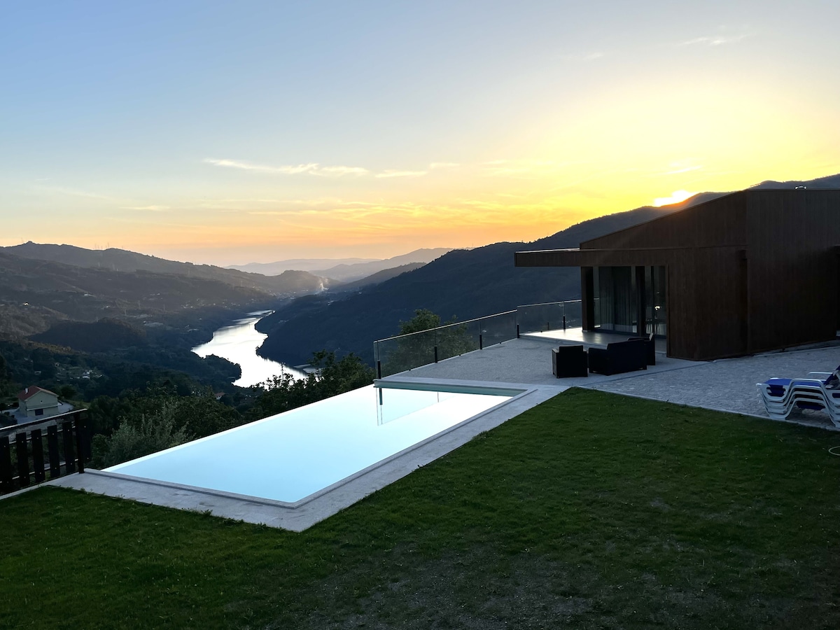 An outdoor area features a modern infinity pool reflecting the vibrant hues of the sunset over rolling hills and the Cávado River. Green lawn surrounds the pool, while a stylish seating arrangement is visible nearby, providing a serene setting for relaxation.