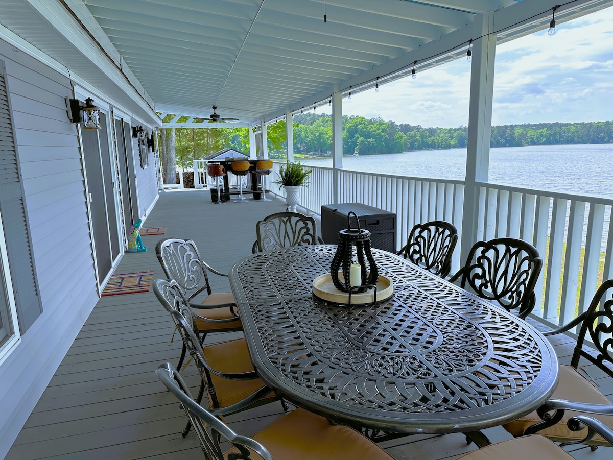 A spacious patio features a large, ornate dining table with seating for eight, surrounded by lush green trees and views of Lake Murray. Decorative string lights hang above, enhancing the tranquil ambiance. A separate area is visible for grilling, offering additional outdoor leisure options.