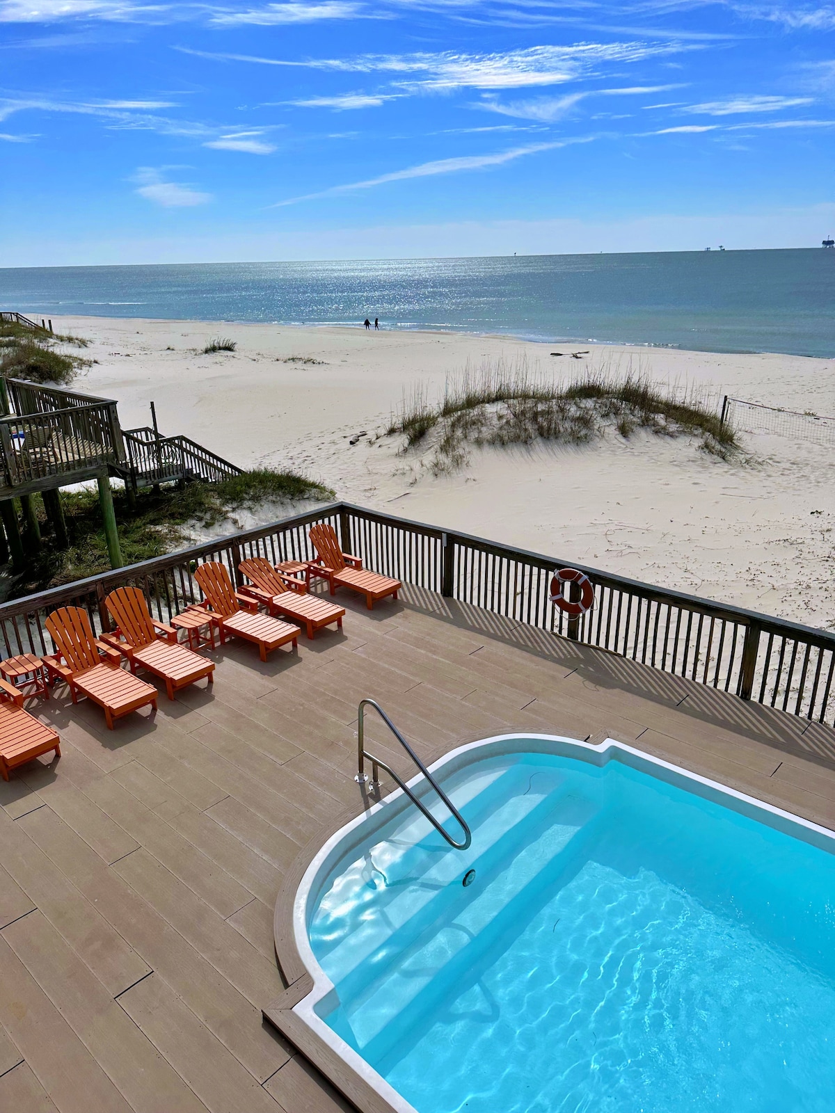 A semi-private pool is shown adjacent to a sandy beach, with the Gulf visible in the background. Six orange lounge chairs are arranged around the pool area, inviting relaxation. Gentle waves glisten under the sun, while sparse dune grass adds to the coastal setting.