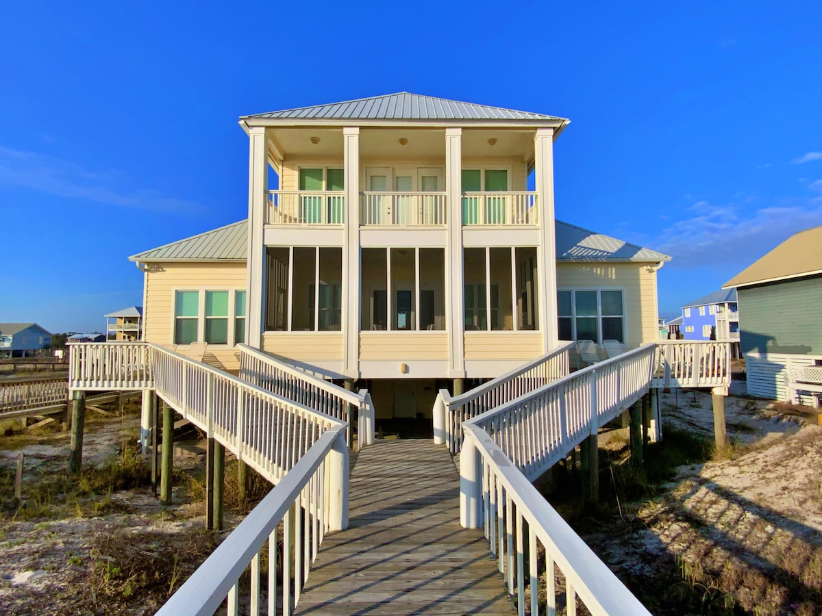 A two-story beachfront home is depicted, featuring a pale exterior and multiple balconies. A wide wooden boardwalk leads to the entrance, flanked by railings. The structure is set against a clear blue sky, with glimpses of nearby beach homes in the background.